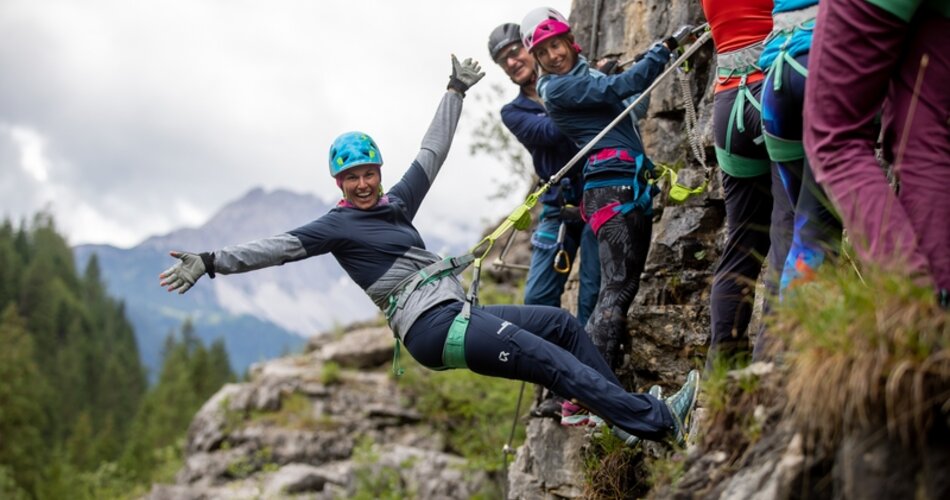 Sechs Leute in einem Klettersteig. Eine Frau hängt sich in den Gurt, streckt die Arme hoch und freut sich. | © AnneKaiser-3274