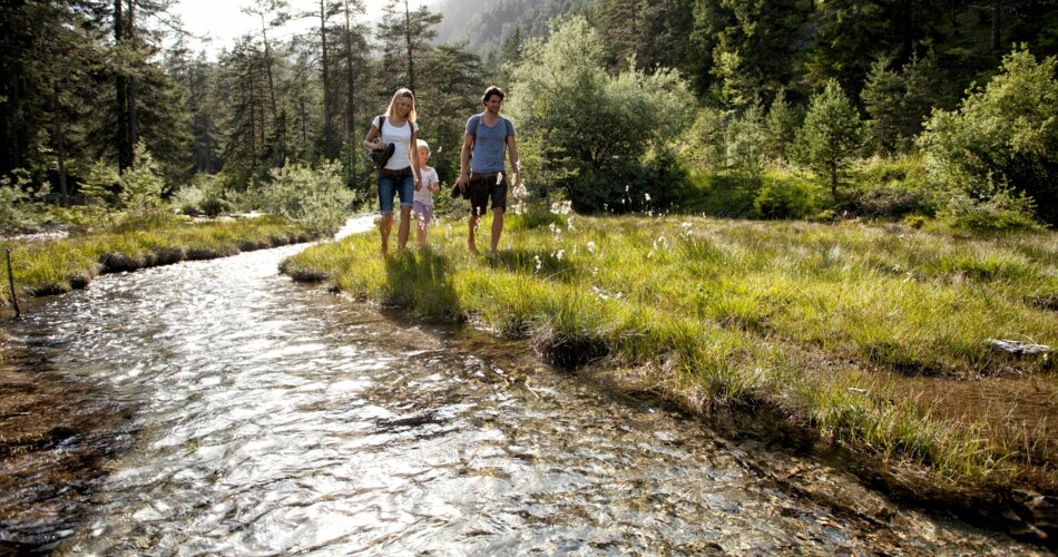 A family of three walks along the Loisach. All around is sparse coniferous forest and green meadows with flowers.  | © Tiroler Zugspitz Arena_U. Wiesmeier