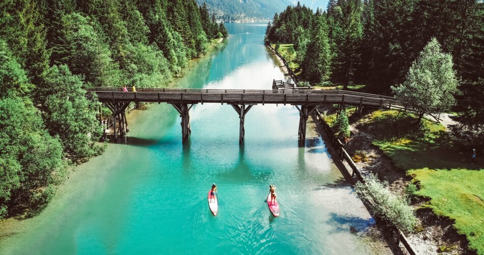 The old bridge at the crossing to Plansee on the turquoise waters of Heiterwanger See. Shortly before the bridge, two people on their stand-up paddle boards. 